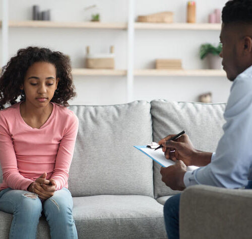 Upset african american curly girl sitting on couch at clinic, having conversation with child psychologist black man, answering questions and looking down. Behavioral disorders in teenagers